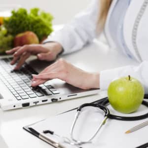 Una dietista scrive al computer portatile. Sulla stessa scrivania sono presenti anche un blocco per appunti, uno stetoscopio e una mela verde in primo piano. Sullo sfondo altra frutta e verdura fresca.