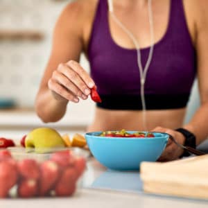 Dettaglio di una giovane donna con un top sportivo e auricolari con filo, che prepara una ciotola di frutta fresca in cucina.