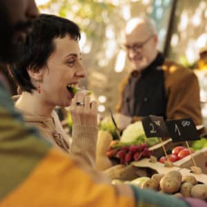 Una donna assaggia uno spicchio di mela mentre è a una bancarella di frutta e verdura, accompagnata da un uomo nero.