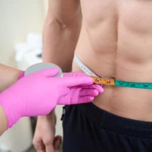 Una dietista con dei guanti monouso rosa fuxia misura con un metro a nastro la circonferenza addominale di un uomo.