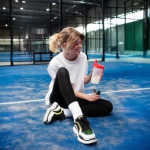 Una giovane donna si riposa seduta per terra durante un allenamento in un campo di tennis o padel al chiuso, mentre beve una bevanda proteica